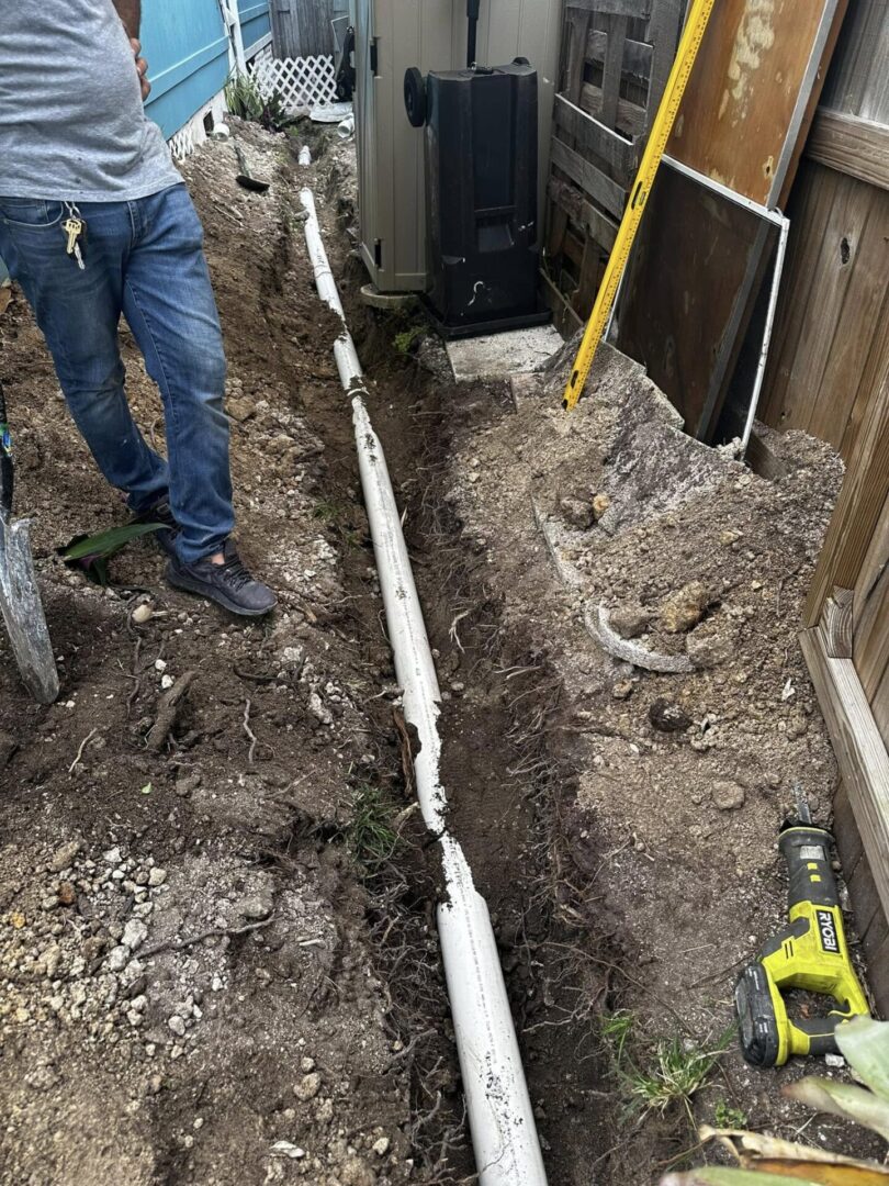 A person stands beside a narrow trench with exposed white pipes and scattered tools. The scene suggests ongoing plumbing or construction work outdoors.