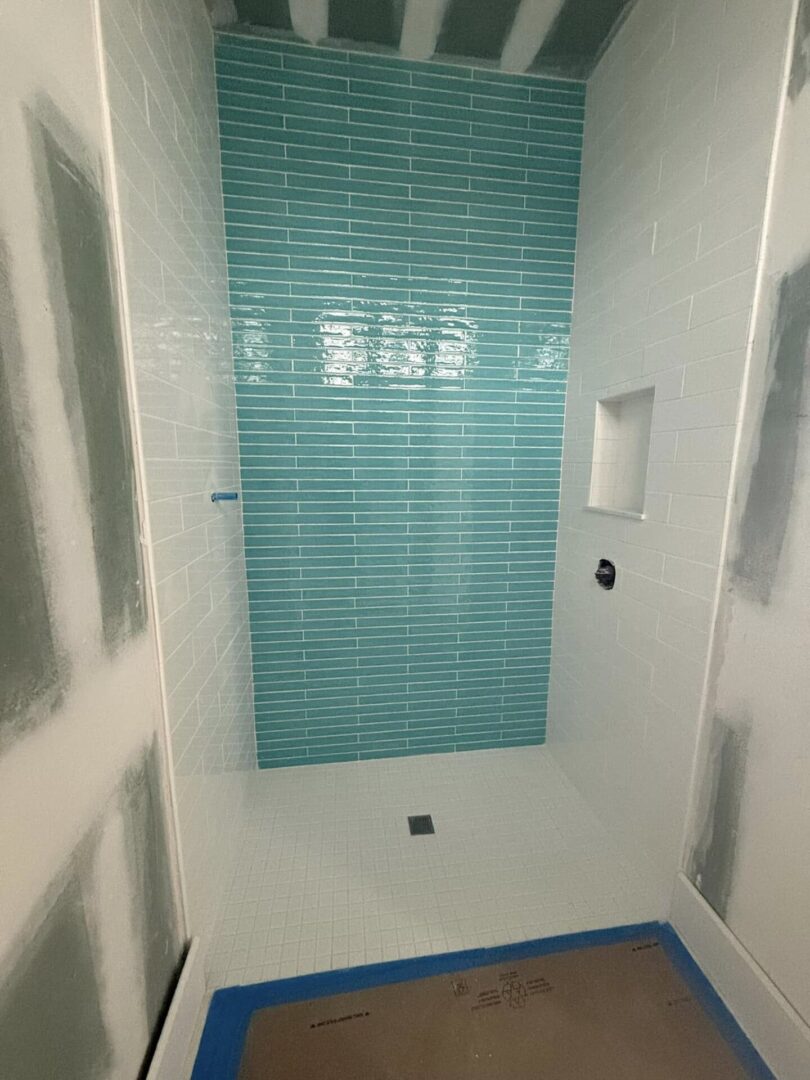 A shower under construction with teal subway tiles, white grout, and a square niche. The floor is white-tiled, with protective cardboard covering the entrance area.