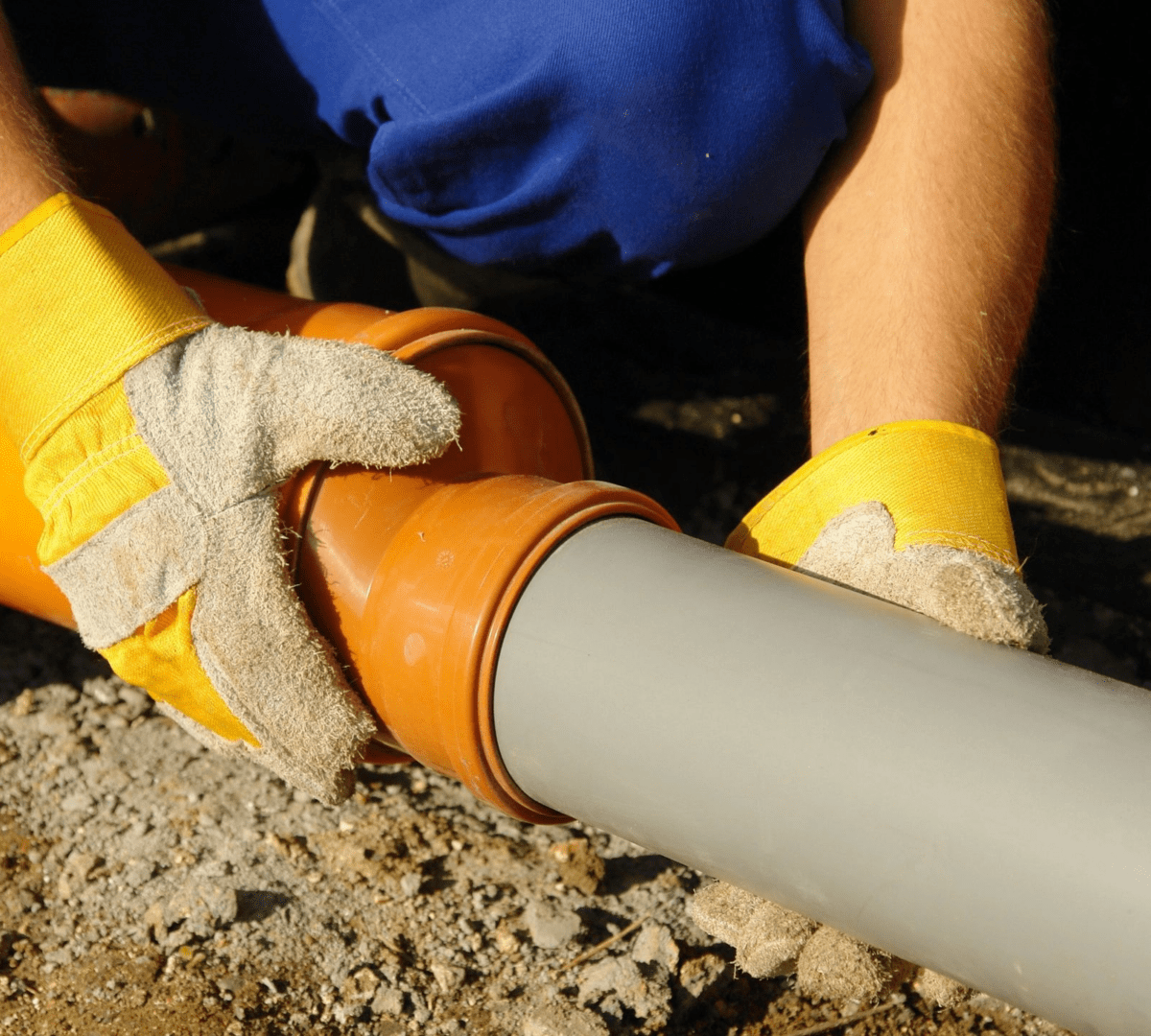 Person with yellow gloves connecting an orange pipe to a gray one on a gravel surface.