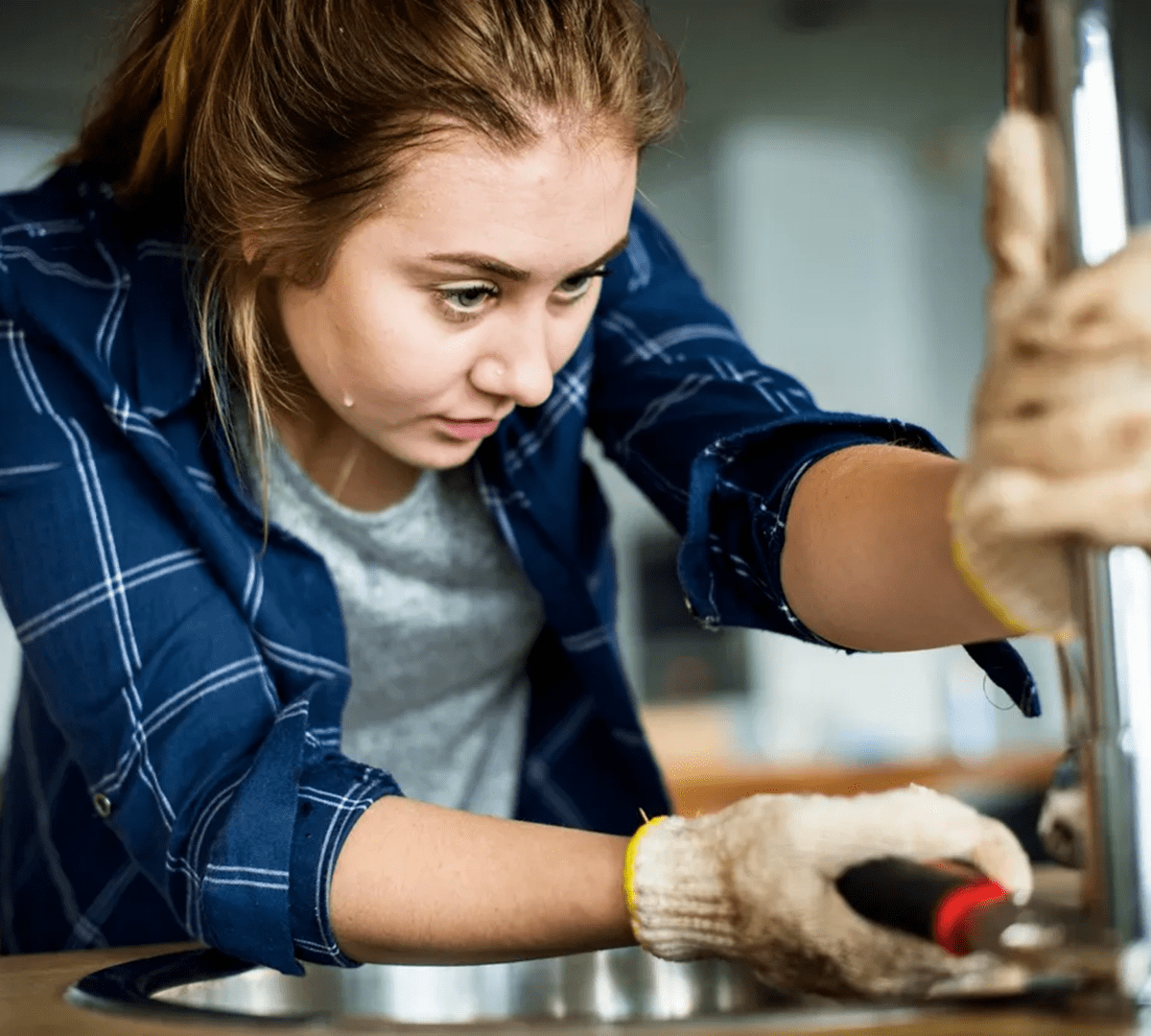 A woman wearing gloves focuses on repairing a kitchen sink faucet, using a wrench.