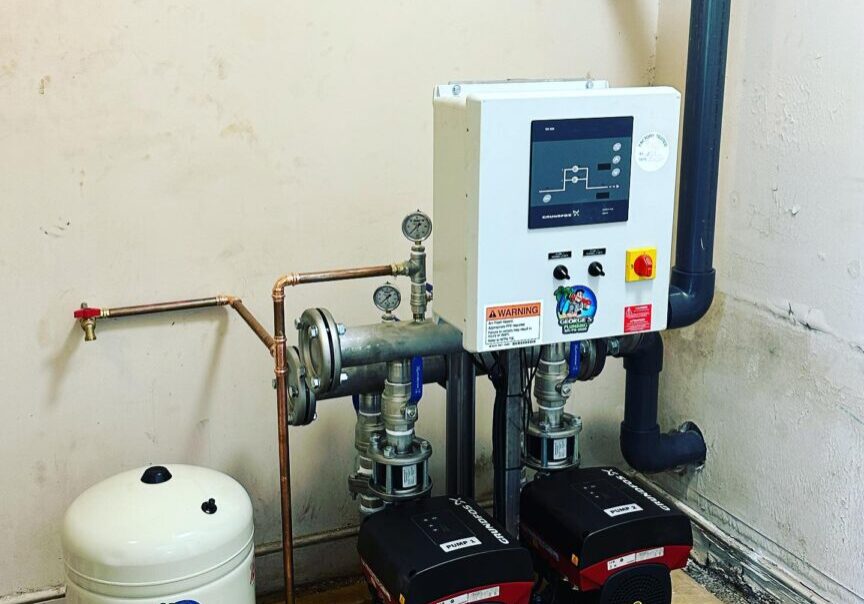 1740473456819_IMAGE-50265_20_287_29 Water pump system with an attached control panel, gauges, two main pumps, piping, and a pressure tank on a concrete floor in a corner.