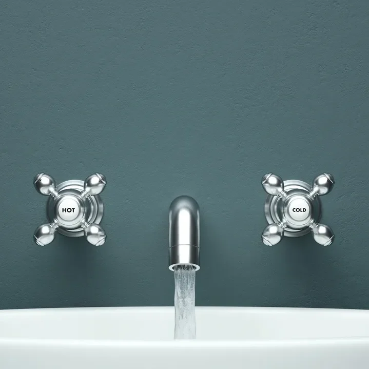 1740473456842_IMAGE-50265_20_289_29 A sink with a chrome faucet and two separate cross-shaped knobs labeled "Hot" and "Cold" against a gray wall. Water is flowing from the faucet into the white basin.