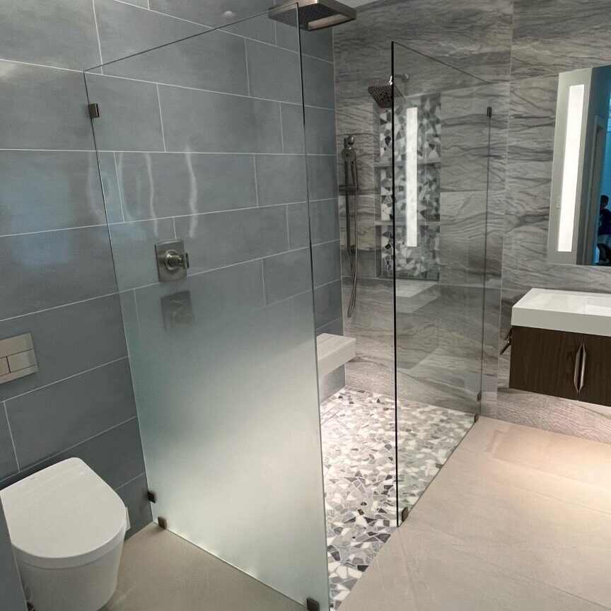 1740473456899_IMAGE-50265_20_289_29 Modern bathroom with a glass-enclosed shower, gray and marble walls, a wall-mounted toilet, and a floating sink with an illuminated mirror. Floor features pebble-like tiles in shower area.