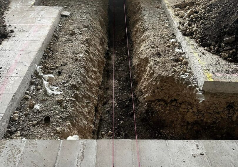 1740473456905_IMAGE-50265_20_2810_29 A trench is dug into the ground, with strings stretched across the top. Surrounding areas show broken tiles and dirt.